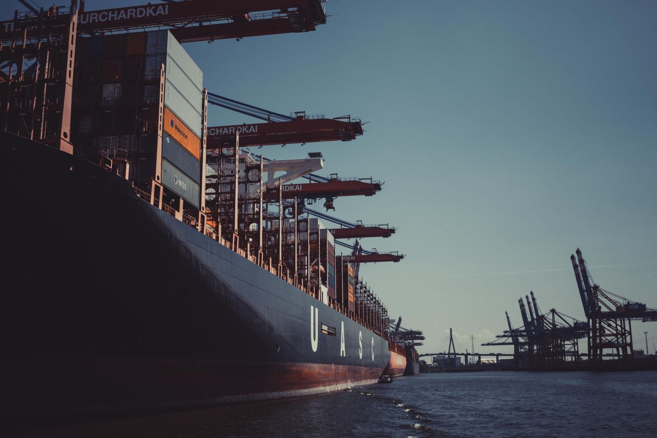 hero-img-02 A large cargo ship docked at Hamburg Harbor, cranes loading containers under a clear sky.