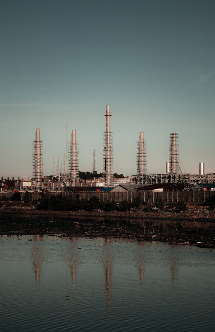 services-01 A refinery with towering smokestacks reflecting in a polluted waterfront, captured during sunset.